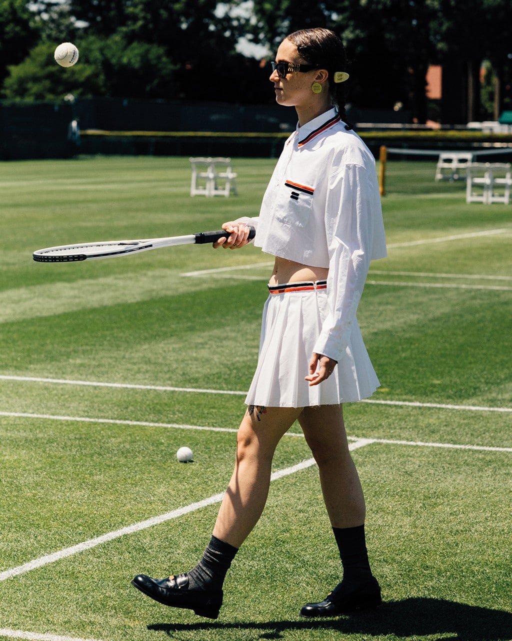 Model wearing a cropped white Miu Miu tennis outfit with loafers and sunglasses on a grass court.